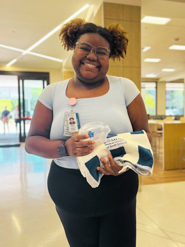 Smiling student holds a MUSC-branded towel and clear piggy bank after winning a campus piggy bank hunt.