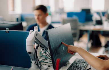 Hand pointing at a computer screen filled with code in a modern office workspace.