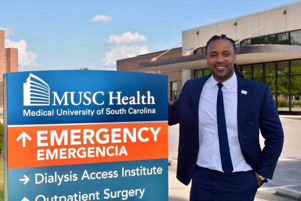 A man wearing a navy suit and tie stands by a sign that says MUSC Health Medical University of South Carolina. Emergency. Emergencia. Dialysis access institute. Outpatient surgery. He is outside with a building behind him.
