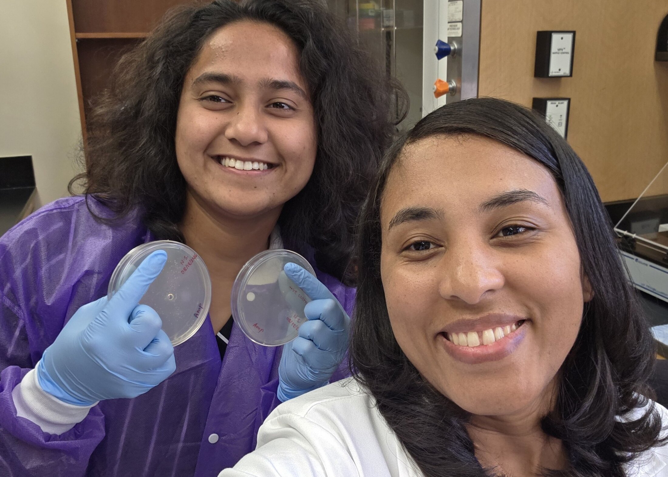 Leidy Diana Caraballo Galva, Ph.D., takes a selfie in her lab with research specialist Niraja Soman, who is holding up two petri dishes.
