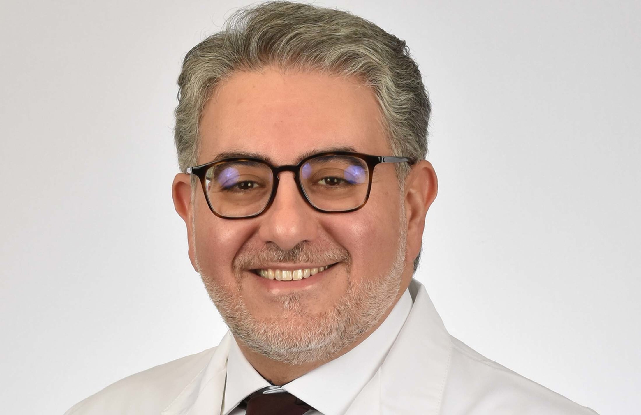 Closeup of a smilng man with gray hear and a beard. He is wearing glasses and a white jacket.