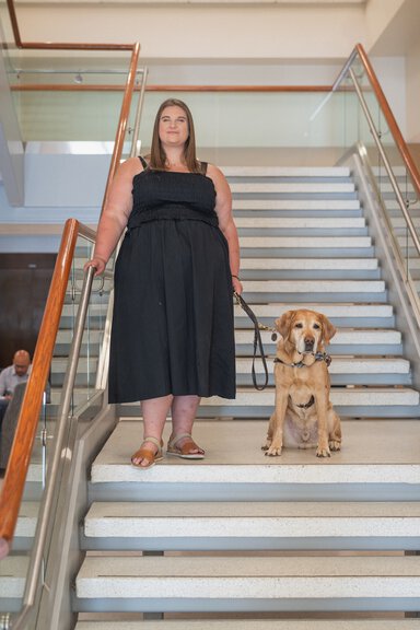 A young woman wearing a black dress is holding the leash of a reddish colored dog who is seated beside her. They are on a stairscase.