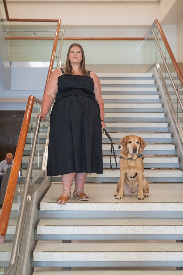 A young woman wearing a black dress is holding the leash of a reddish colored dog who is seated beside her. They are on a stairscase.