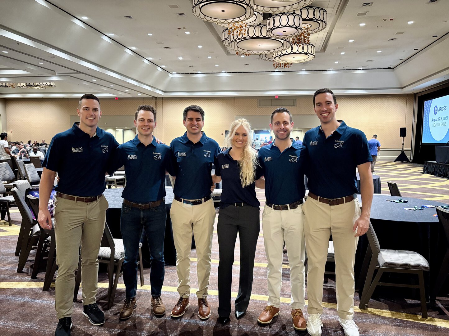 Six people wearing matching navy MUSC-branded polo shirts pose together in a large conference ballroom during a professional meeting. They are smiling with their arms around one another, standing in front of banquet tables and chairs, with a presentation stage and conference screen visible in the background.