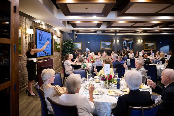 A woman speaks at a podium in a banquet hall with elegantly set tables. Attendees, mostly older adults, listen attentively. A screen displays 