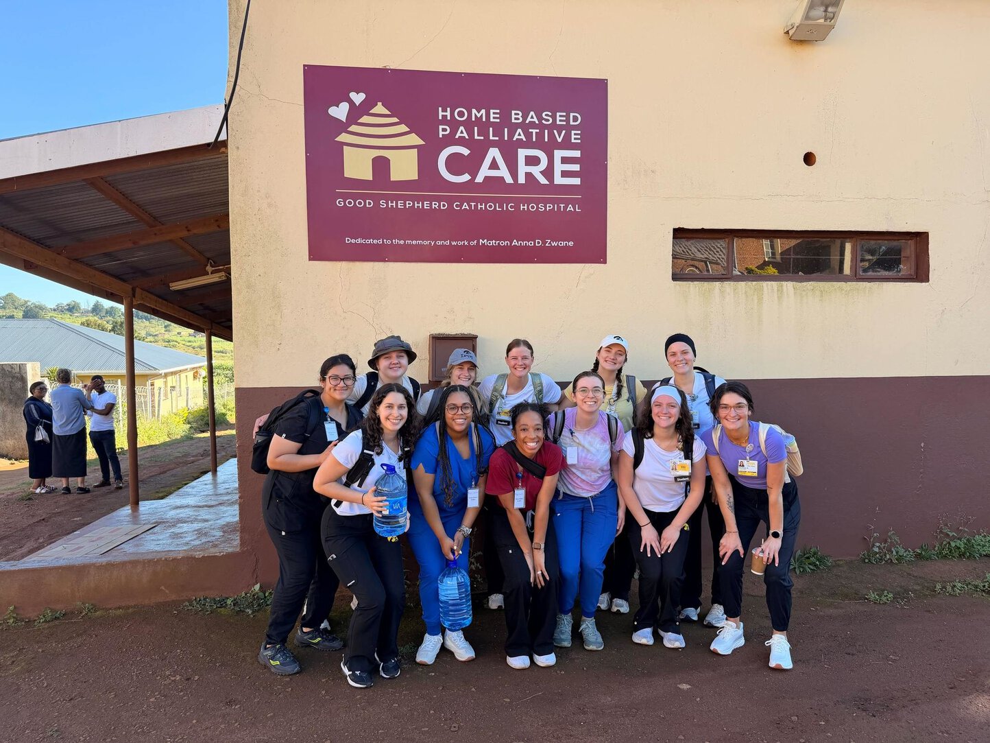 A group of approximately twelve nursing students and faculty pose together outside a building with a maroon sign reading Home Based Palliative Care – Good Shepherd Catholic Hospital, dedicated to Matron Anna D. Zwane.