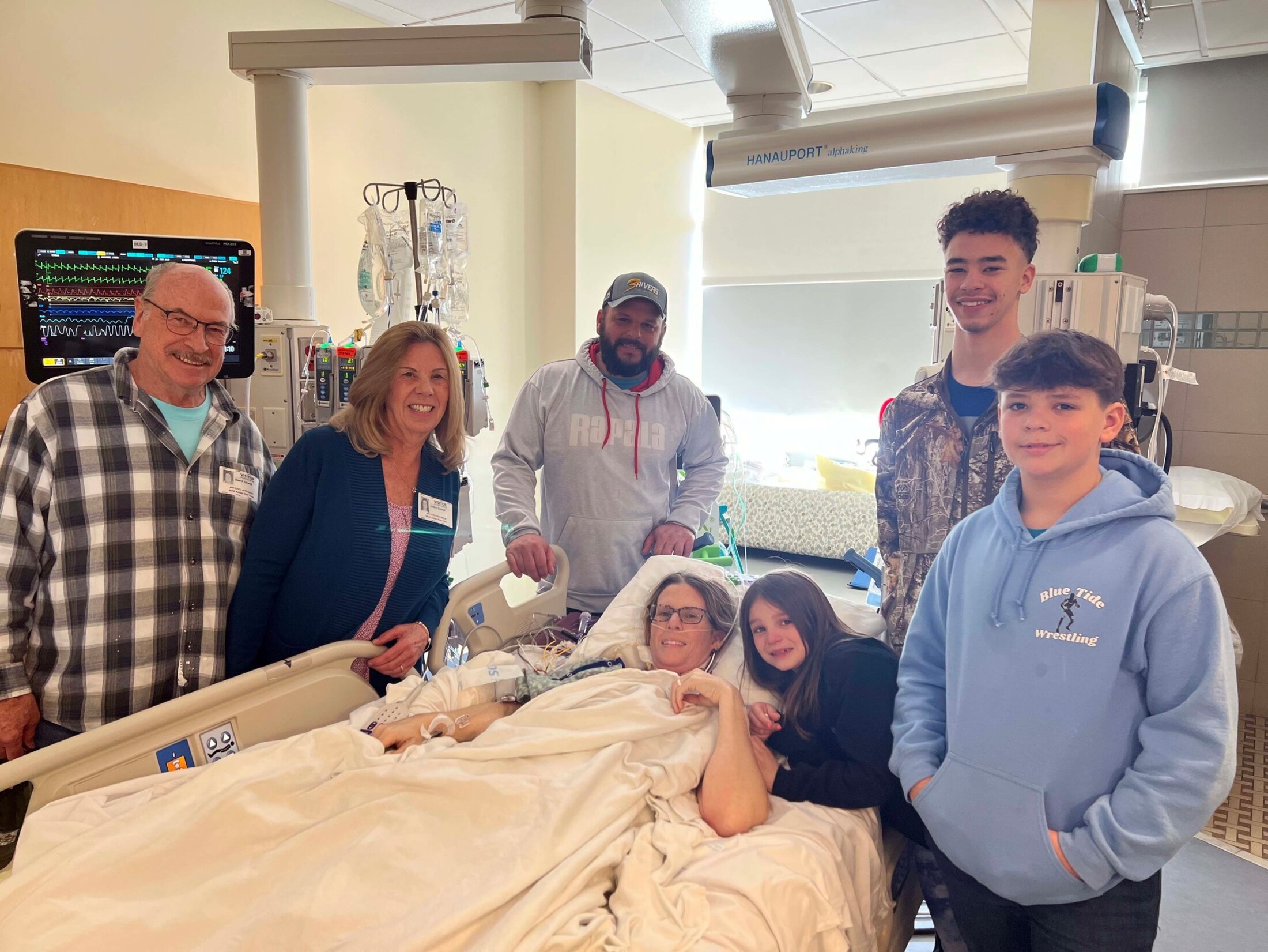 A woman is lying in a hospital bed surrounded by two older people on the left, a bearded man center and three children on the right.