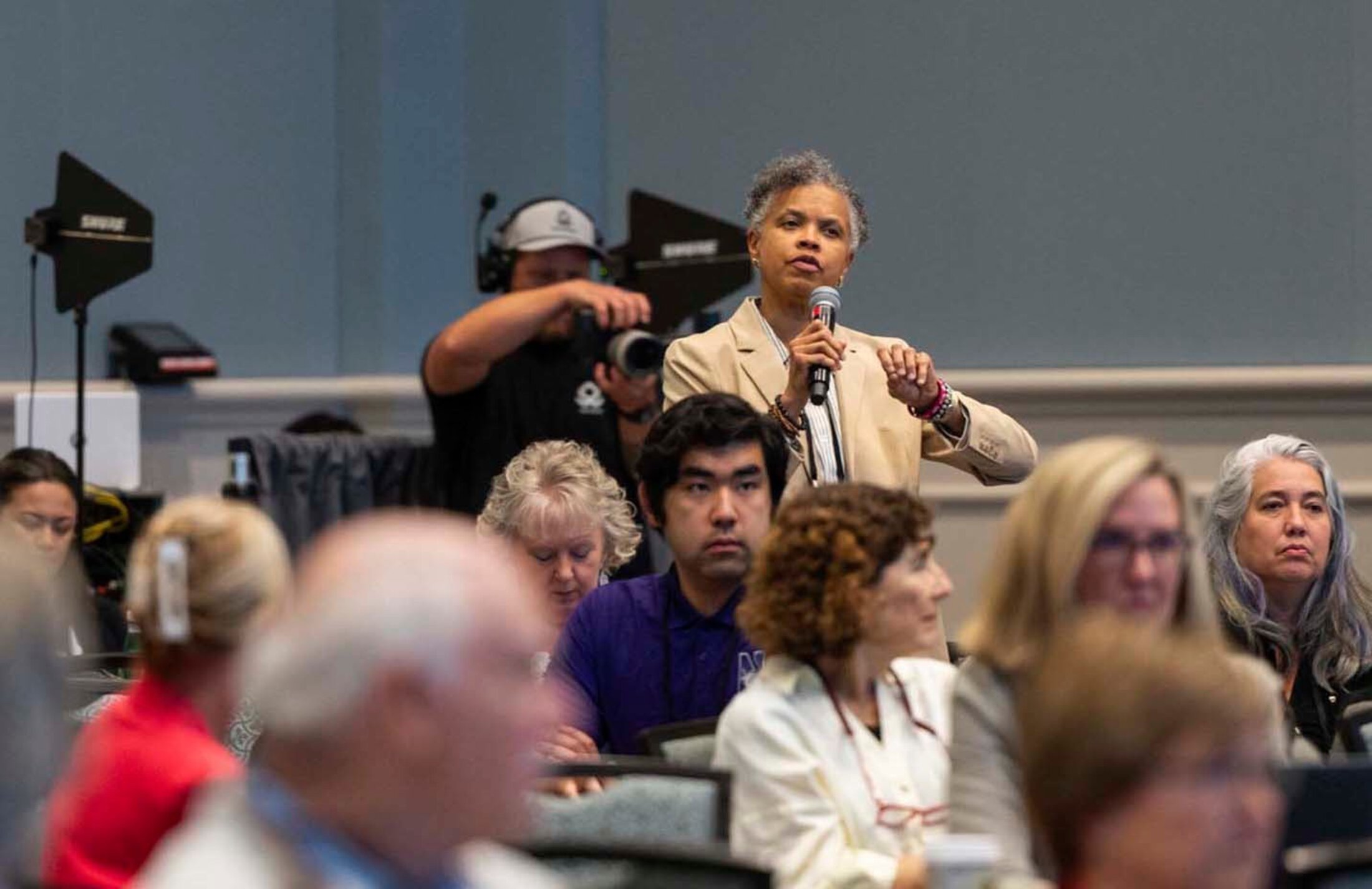 A woman stands in an audience holding a microphone. Sheh is wearing a tan blazer. Her head is cocked as she speaks. A photographer is behind her. She's surrounded by seated adults.