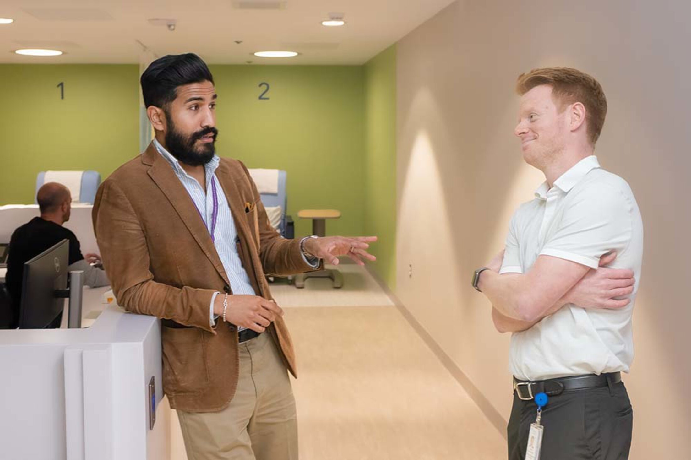 Two men stand in a hallway talking. The one on the left is gesturing. The one on the right has his arms crossed and is smiling.