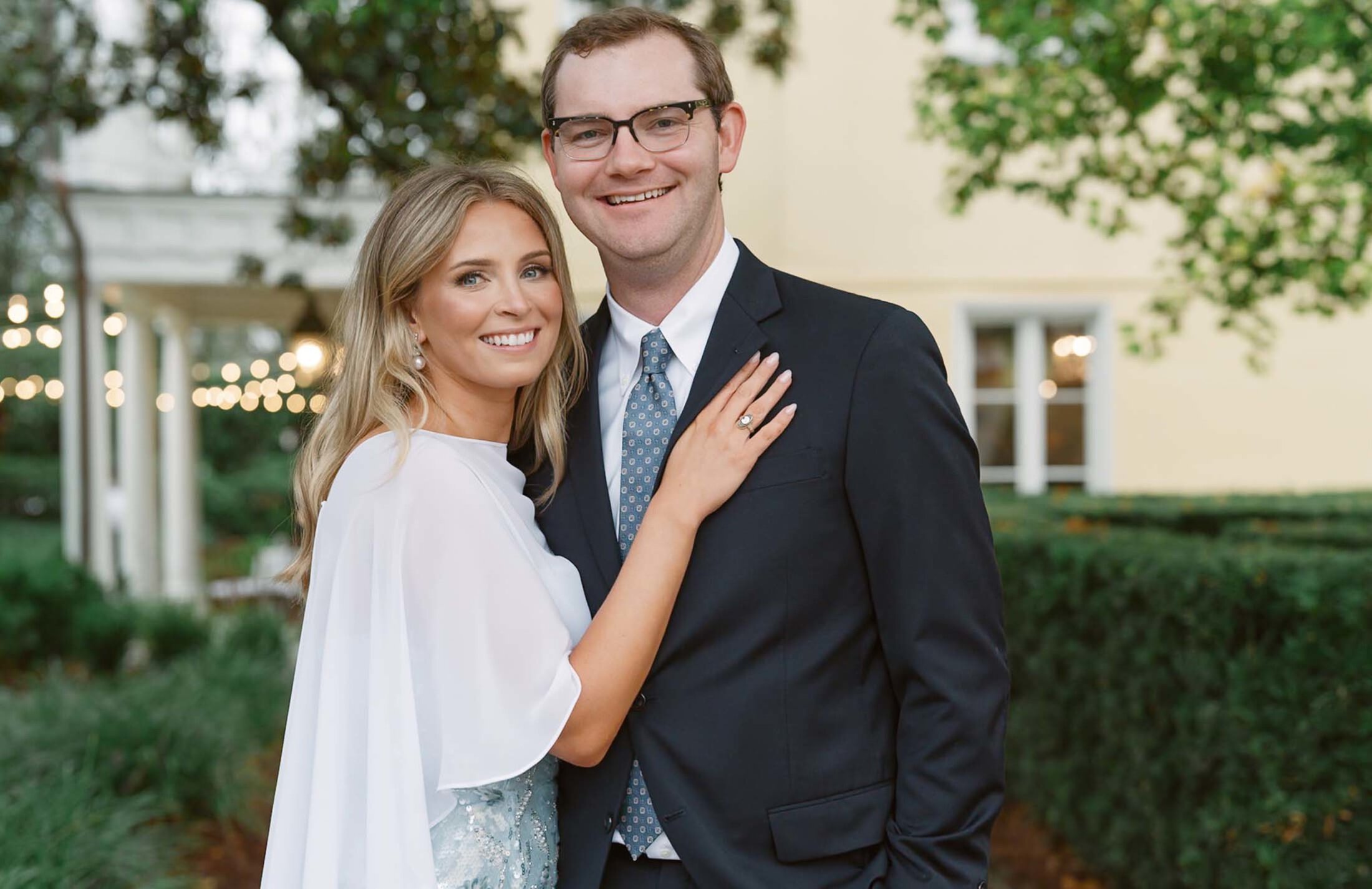 A wedding portrait of a woman and man standing outside with trees and a building behind them. She is wearing a white sheer cape with a sequined dress. Her arm is on his shoulder. He is in a dark suit and glasses. Both are smiling.
