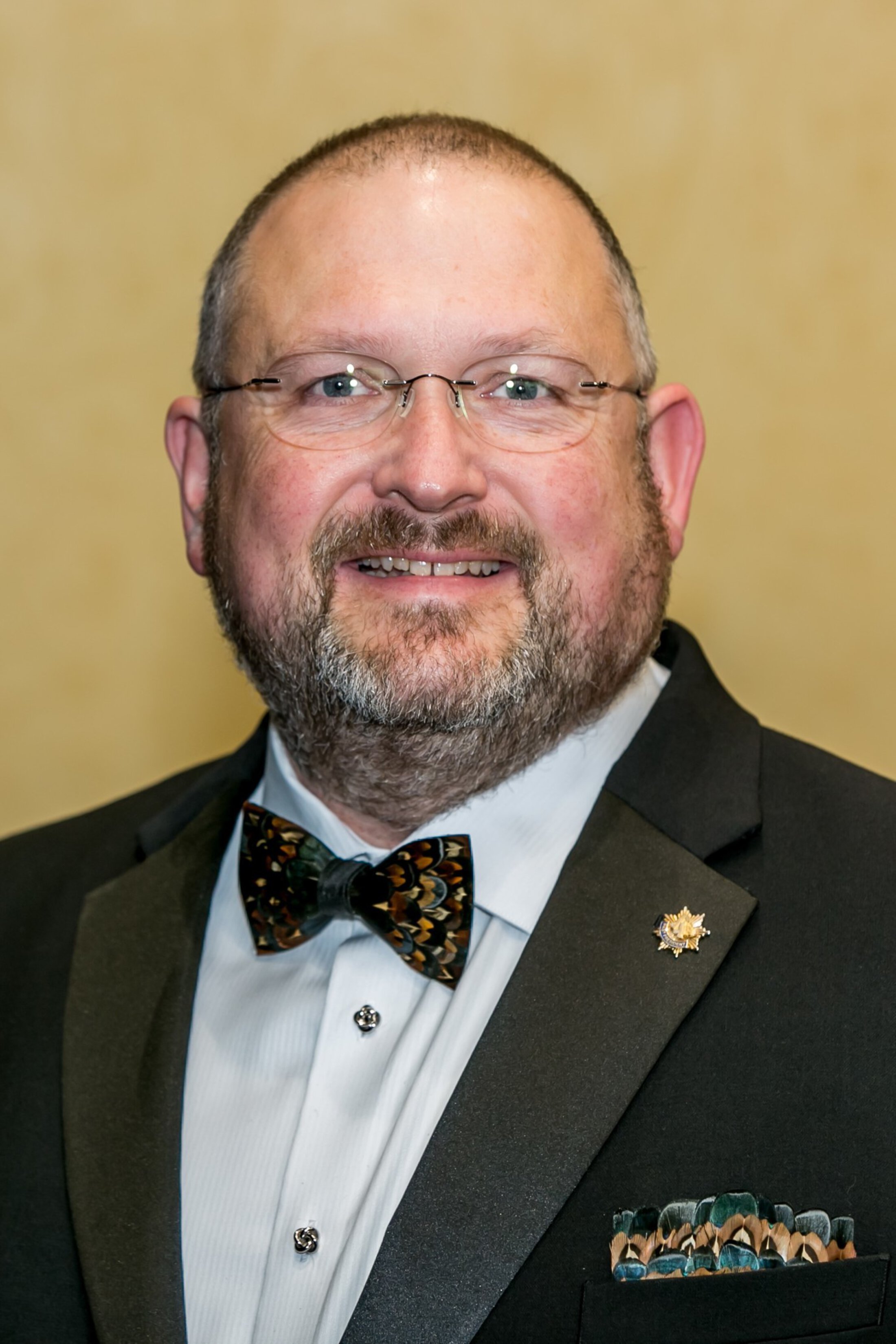 Man with glasses and a neatly trimmed beard smiling at the camera, dressed in a black tuxedo with a patterned bow tie and matching pocket square against a neutral background.