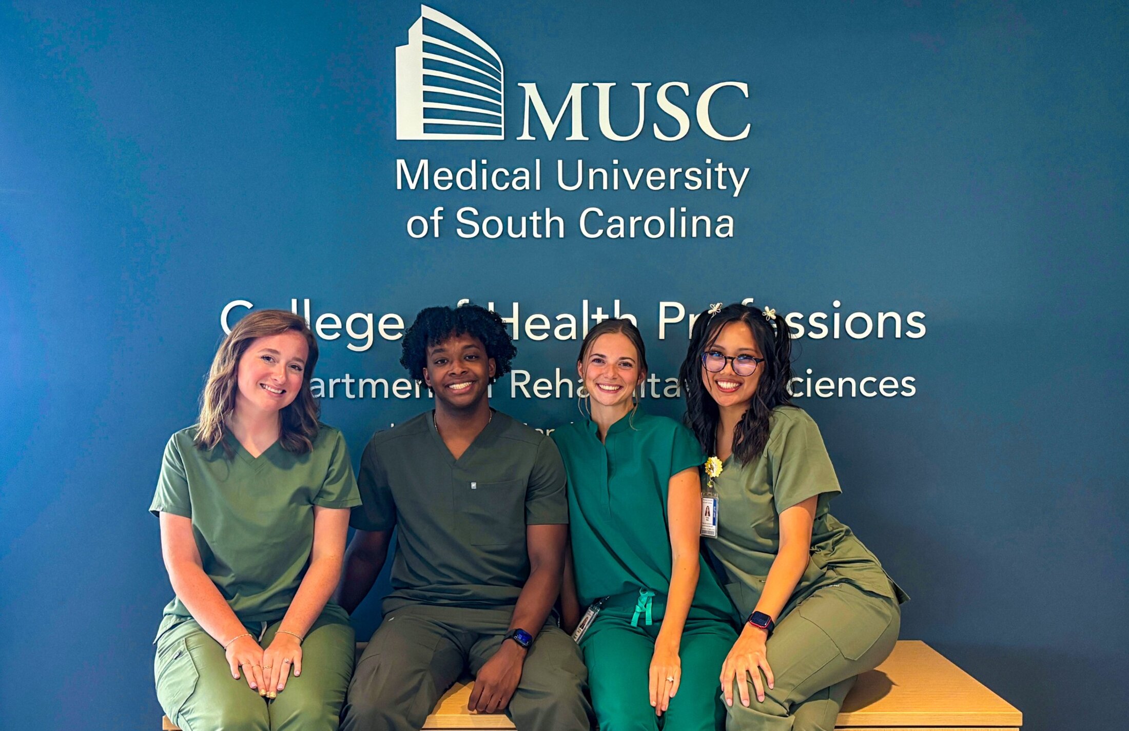 Four young adult students in green scrubs sit together beneath a wall displaying the MUSC logo and College of Health Professions Department of Rehabilitation Sciences signage.