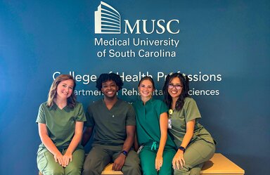 Four young adult students in green scrubs sit together beneath a wall displaying the MUSC logo and College of Health Professions Department of Rehabilitation Sciences signage.