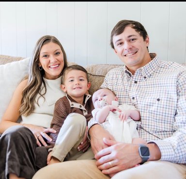 A family of four sitting together on a couch, including an adult man, woman, a young boy, and an infant.