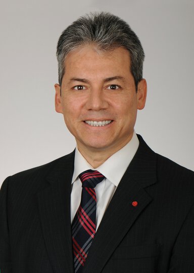 Headshot of a smiling man with gray short hair. He is wearing a white shirt, a black, red and white tie and a black sport coat with a small red pin.
