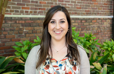 A woman posing for her portrait stands outside in front of a brick wall and folliage and smiles at the camera.