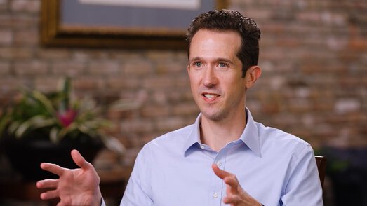 A seated man speaks to the camera in front of a brick wall