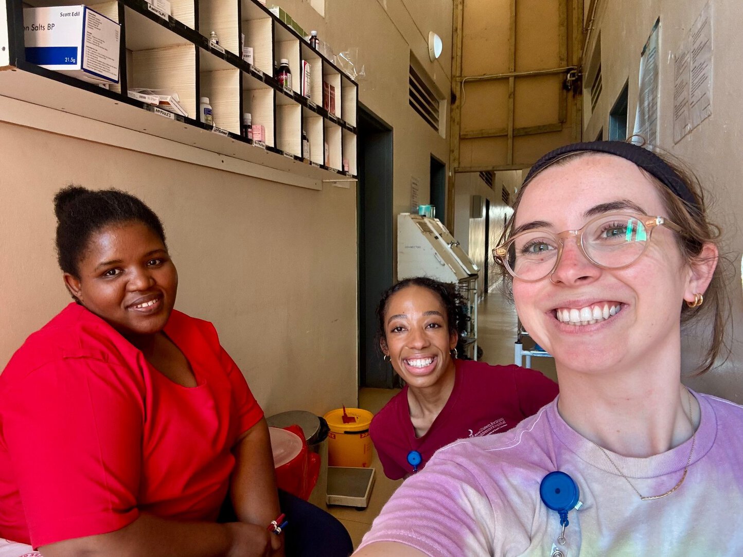Three women smile for a selfie inside a medical facility. Behind them are wall-mounted shelves stocked with medications and supplies. Two of the women wear lanyards with ID badges.