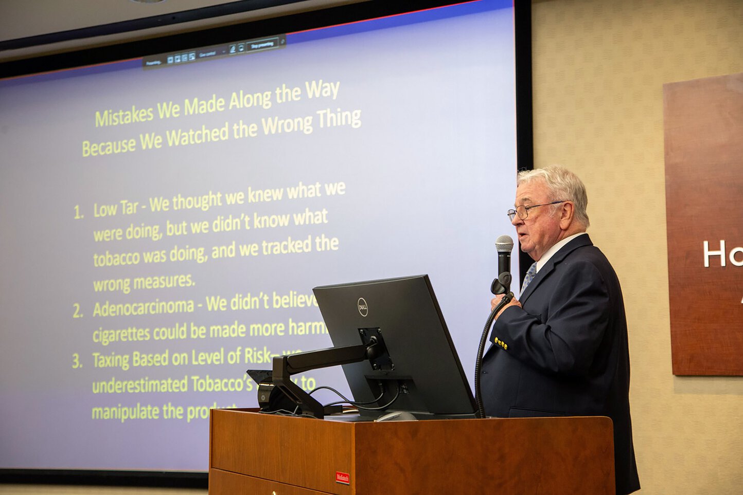 A man speaks at a podium with a large screen showing slides behind him.