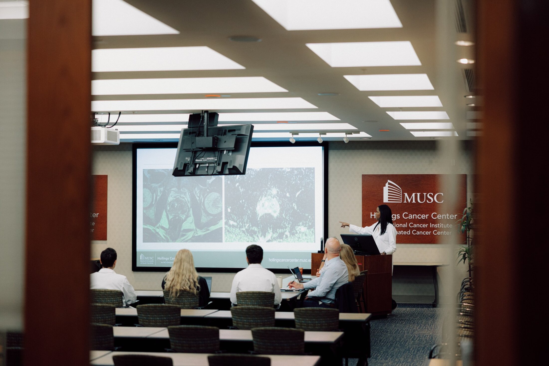 A physician at a podium points to scans displayed on a large screen for a group to discuss.
