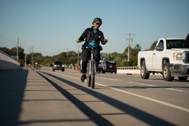 A woman smiles as she bicycles on a bike path over a bridge.