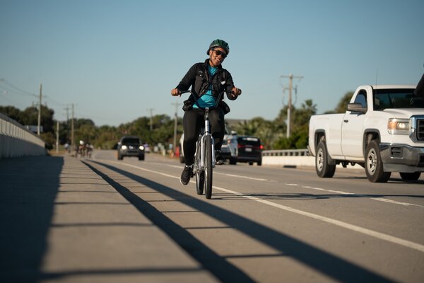 A woman smiles as she bicycles on a bike path over a bridge.