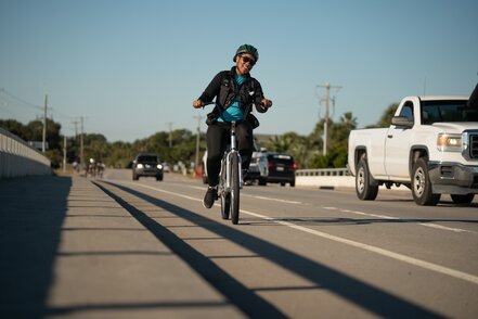 A woman smiles as she bicycles on a bike path over a bridge.