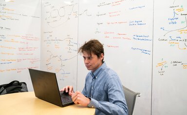 A man works at a laptop in a room where the walls are whiteboards covered in scribbles outlining cellular biology and pathways.