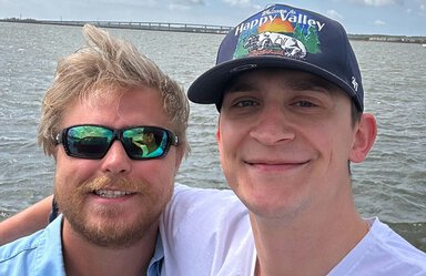Two young men with water behind them. The man on the left has blond hair and a beard. he's wearing sunglasses and a blue shirt. The one on the right is wearing a baseball cap that says Welcome to Happy Valley. He has dark eyebrows and is wearing a white t-shirt.
