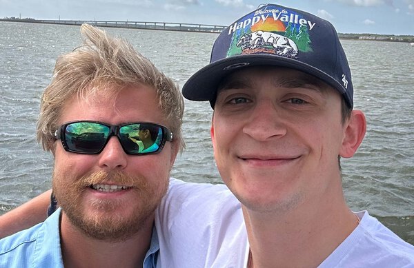 Two young men with water behind them. The man on the left has blond hair and a beard. he's wearing sunglasses and a blue shirt. The one on the right is wearing a baseball cap that says Welcome to Happy Valley. He has dark eyebrows and is wearing a white t-shirt.