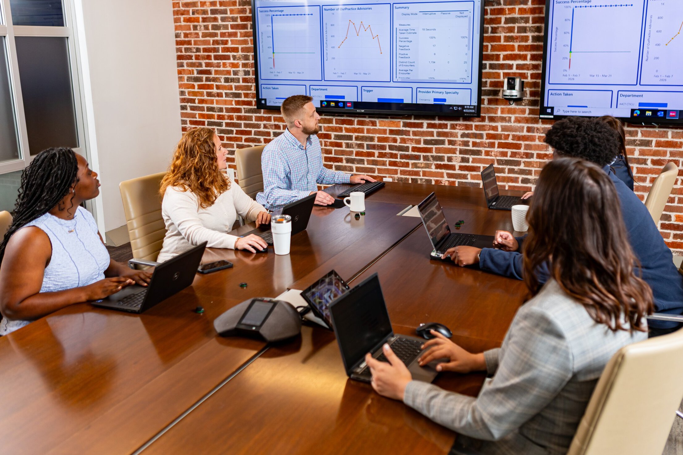Several men and women are gathered around a conference table reviewing data on large screens.
