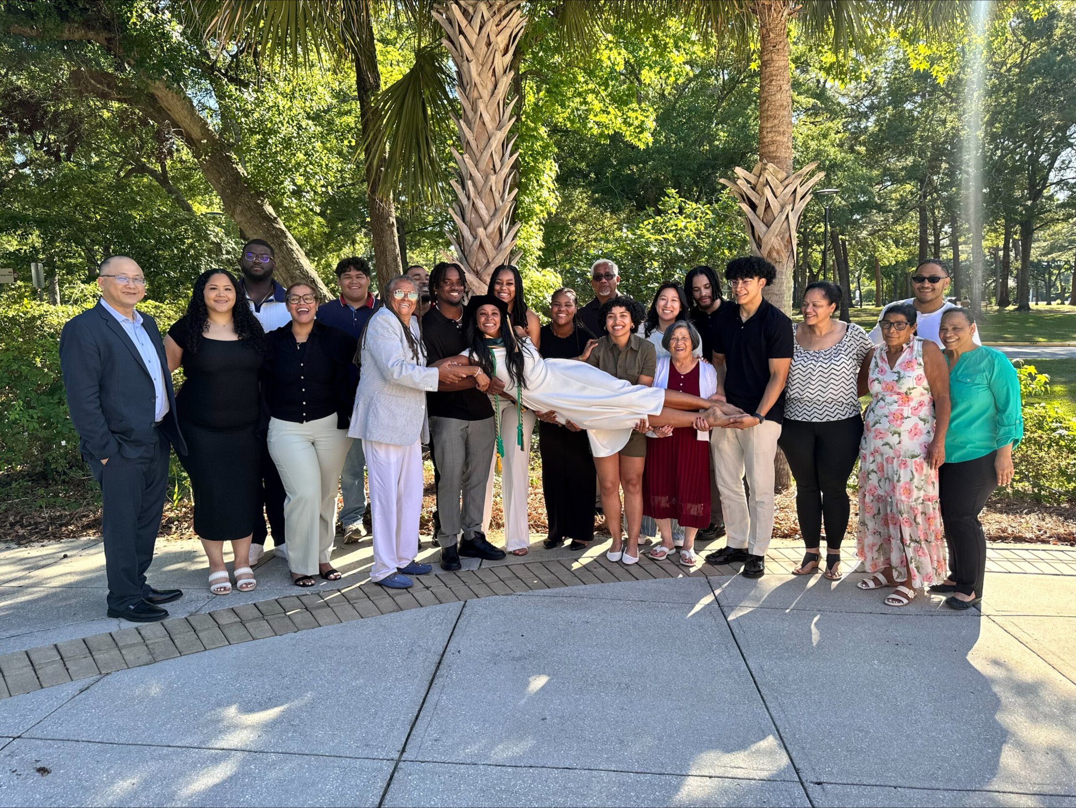 Large family group gathered outdoors beneath palm trees, smiling as they hold a graduate in a white dress horizontally in celebration. The group spans multiple generations and poses on a sunny walkway surrounded by lush greenery.