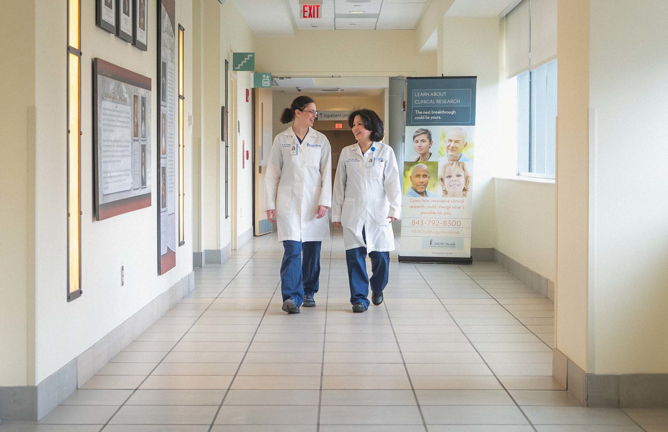 Two women wearing white coats and blue scrubs walk down a hallway. There is a banner behind them that says learn about clinical research.