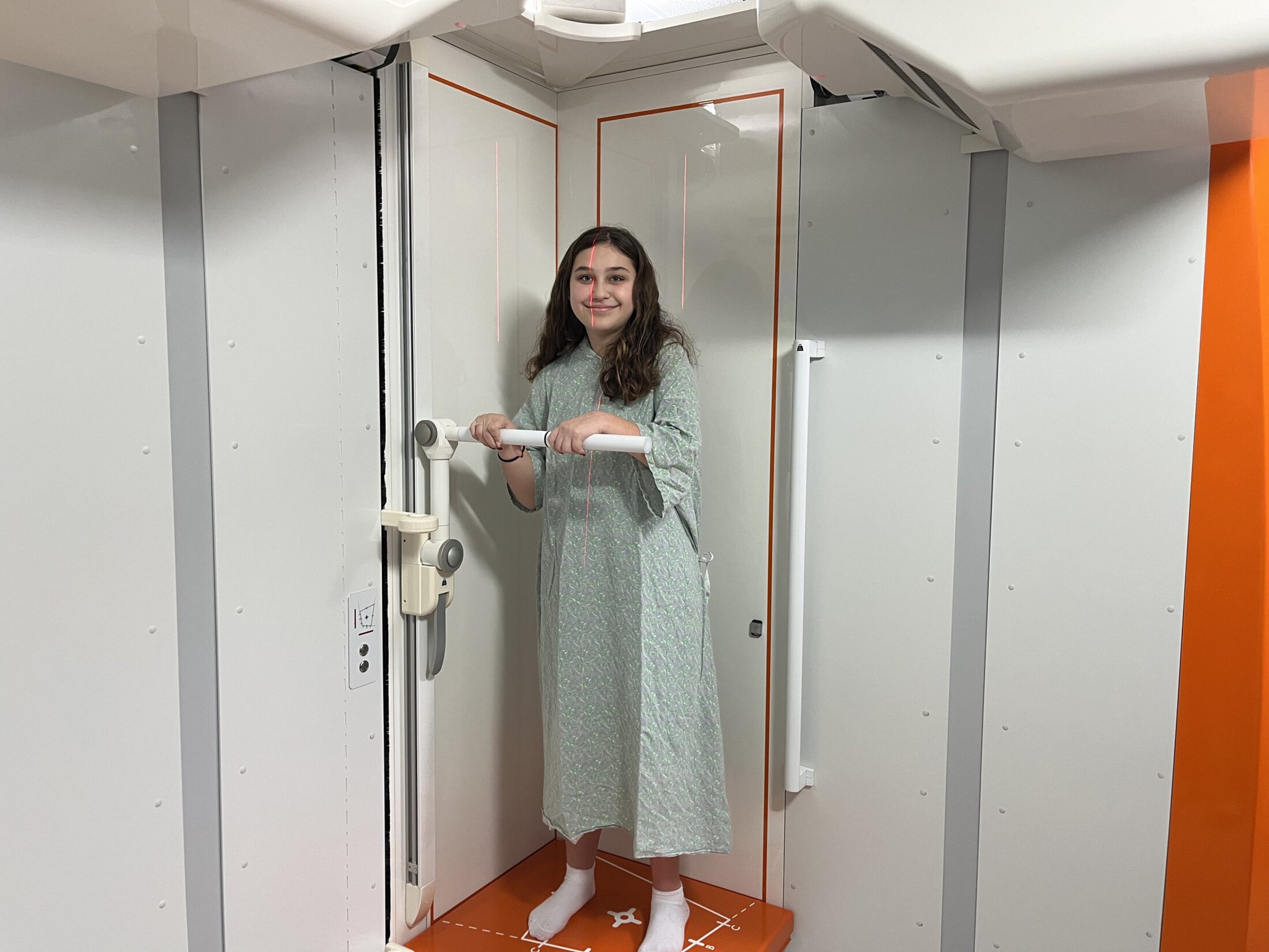 A young woman in a hospital gown stands inside an EOS imaging machine, holding a horizontal handle bar while laser alignment lines are visible across her body. The machine's interior is white with orange trim.