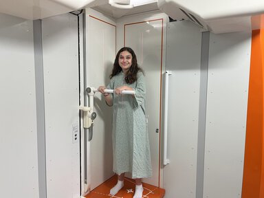 A young woman in a hospital gown stands inside an EOS imaging machine, holding a horizontal handle bar while laser alignment lines are visible across her body. The machine's interior is white with orange trim.