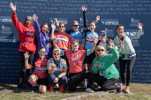 a large group of riders stand in front of a Lowvelo backdrop cheering and raising their arms in the air