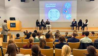 Graduate student with short hair and glasses presents research at a podium while undergraduate students listen attentively in an auditorium.