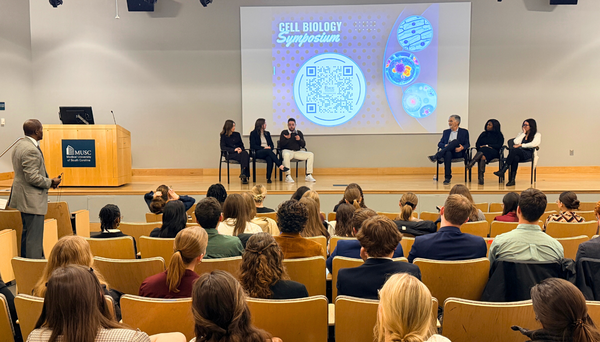 Graduate student with short hair and glasses presents research at a podium while undergraduate students listen attentively in an auditorium.