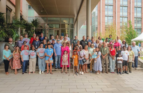 Three rows of people standing on stairs with the glass entrance to a building behind them. Some are wearing t-shirts that say ECMO reunion 2026. Most are smiling.