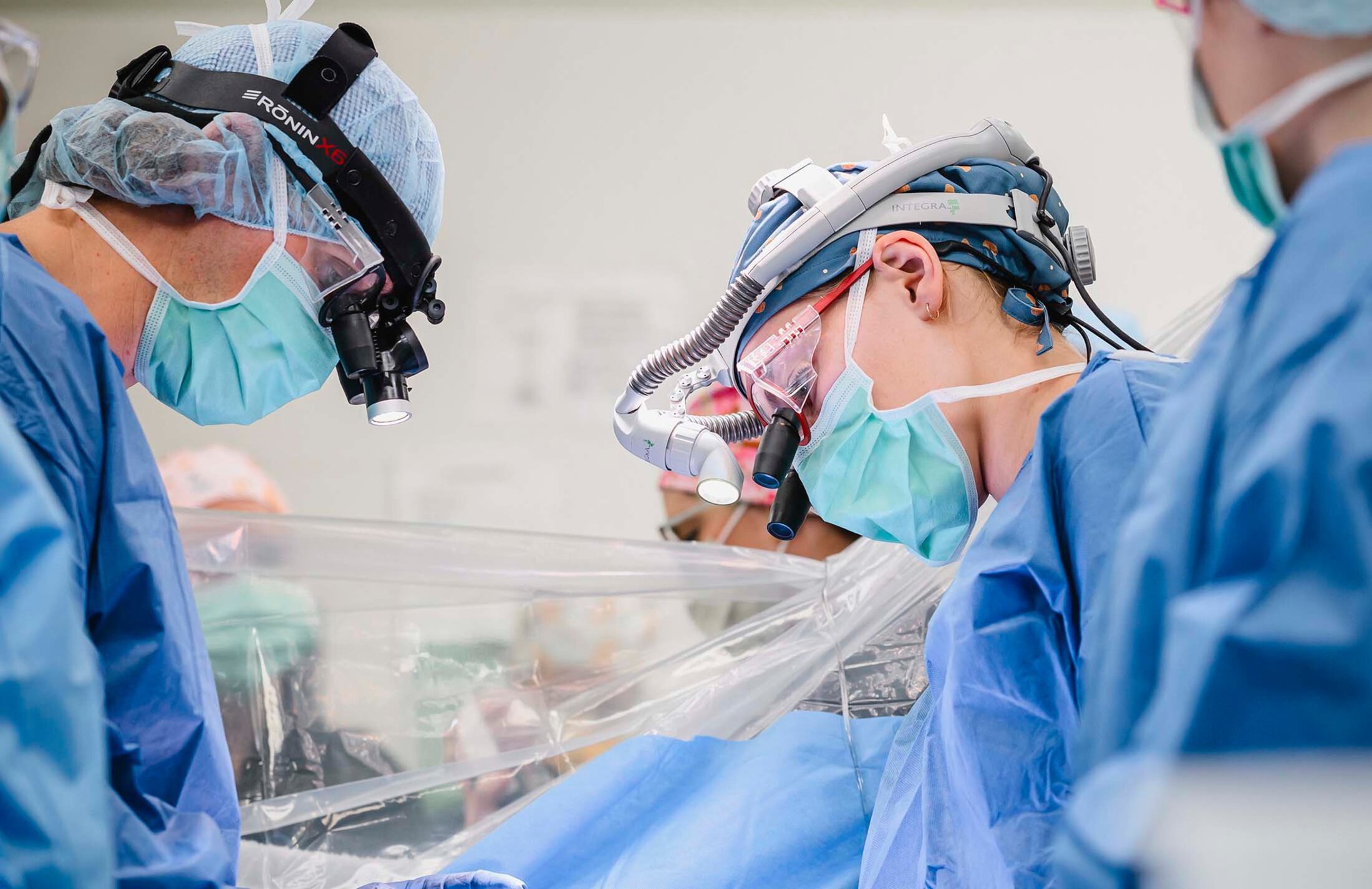 Four people wearing blue hospital scrubs in an operating room. The two featured people are wearing special equipment on their heads - special lenses and one is also wearing a light.