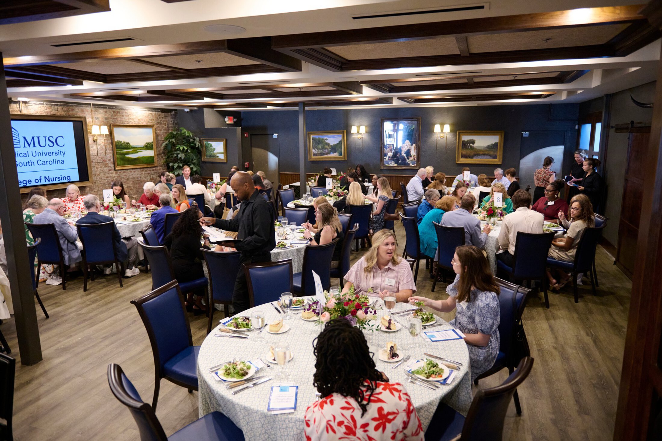 A woman speaks at a podium in a banquet hall with elegantly set tables. Attendees, mostly older adults, listen attentively. A screen displays 