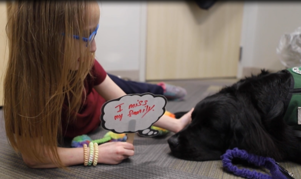 A child patient holding a I miss my family hand drawn sign and petting a black lab dog