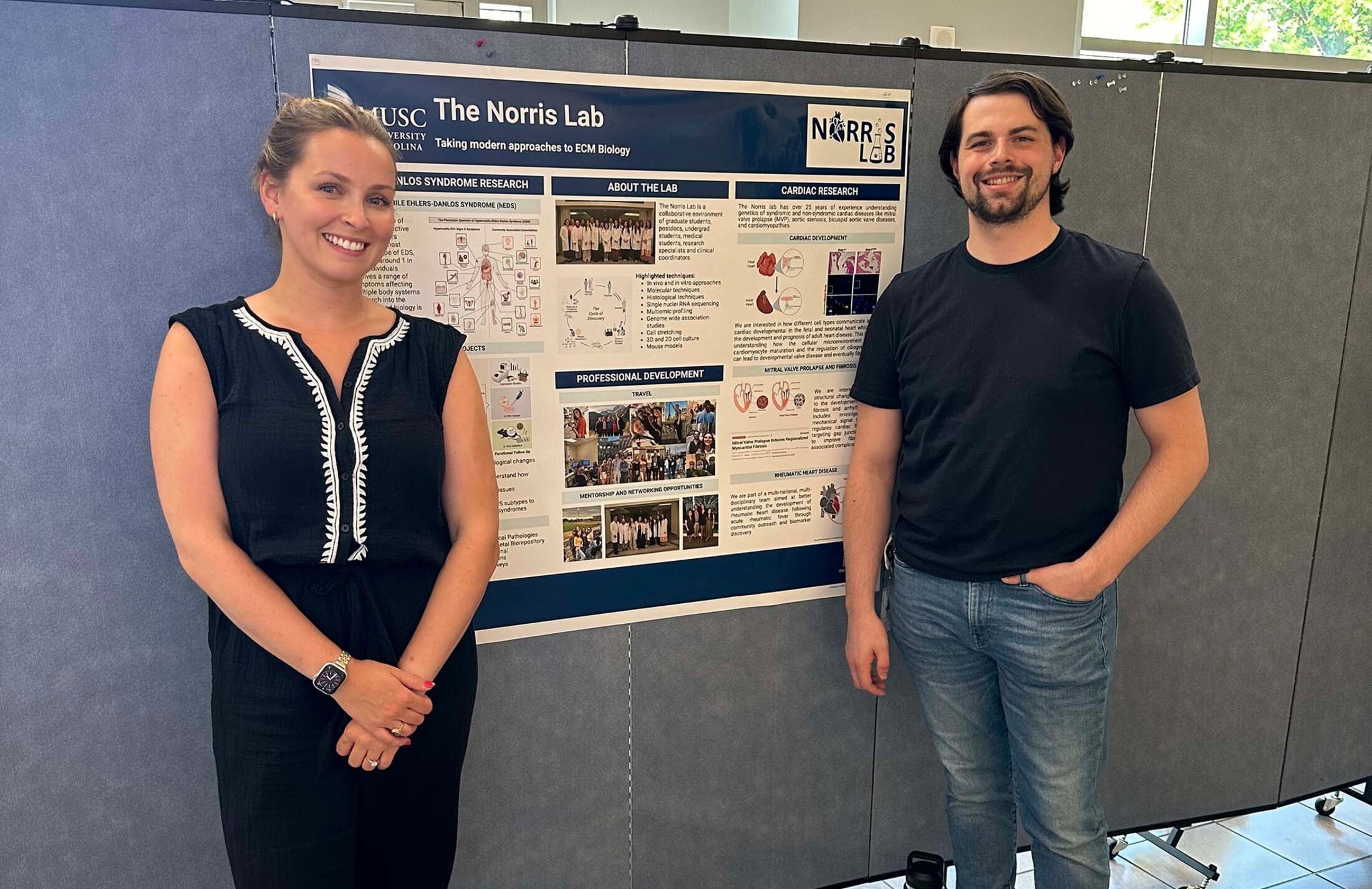 Two people stand beside a poster that says The Norris Lab. Taking modern approaches to ECM biology. The woman on the left has her hair pulled back and is wearing a navy and white outfit. The man on the right has a goatee and is wearing a dark shirt with jeans. Both are smiling.