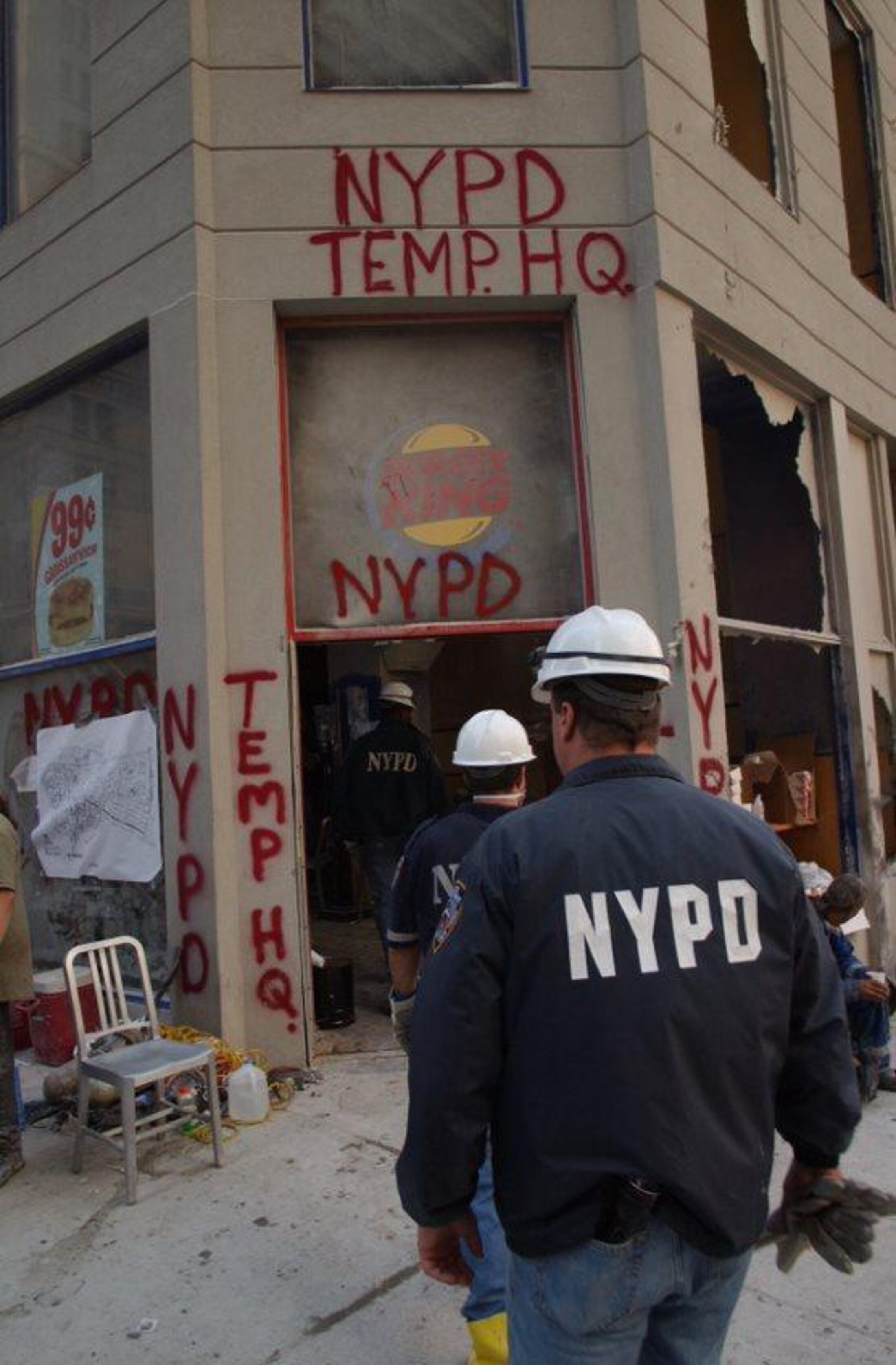 Outside of a building has the words NYPD Temp. HQ (headquarters) spraypainted on it. Two men wearing NYPD jackets and construction helmets are walking toward it.