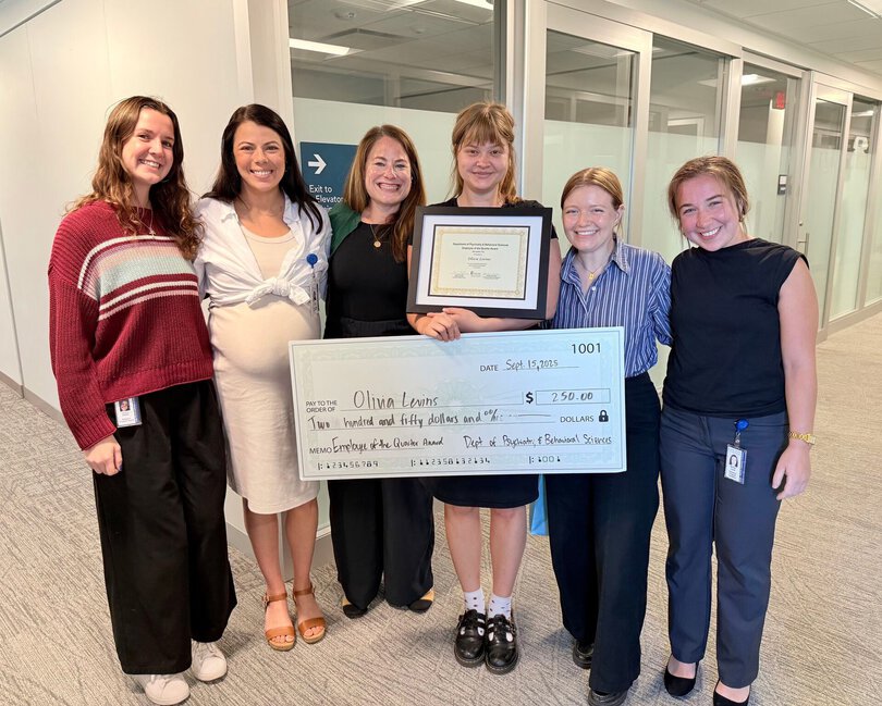 A group of women stands together with the woman in the middle holding a framed certificate and an oversized novelty check.
