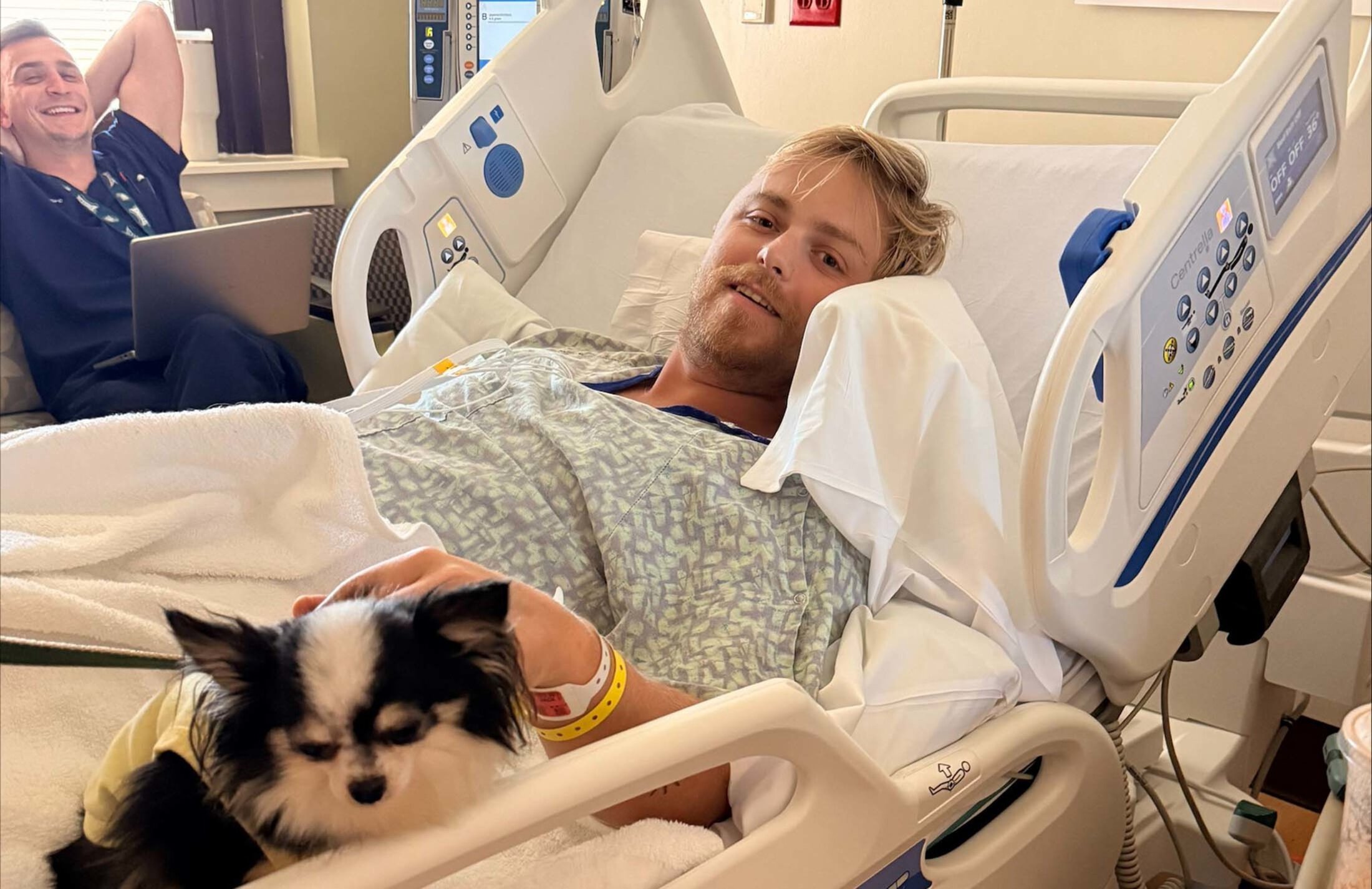 A young man with blond hair and a beard is lying in a hospital bed He is wearing a gown and has his left hand on the back of a small black and white dog. On the left side is another man. He's wearing blue hospital scrubs and has his hands behind his head. He has a big smile.