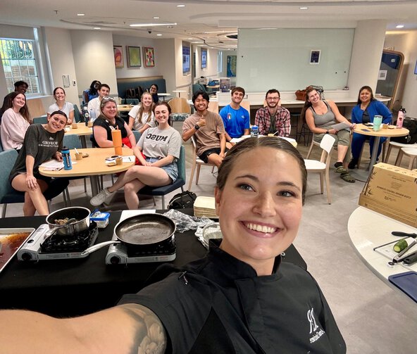 Smiling chef takes a selfie with a group of students seated around tables during an interactive cooking class.