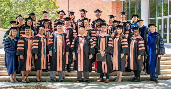 A class of nearly 30 men and women pose in caps and gowns outdoors following a graduation ceremony.