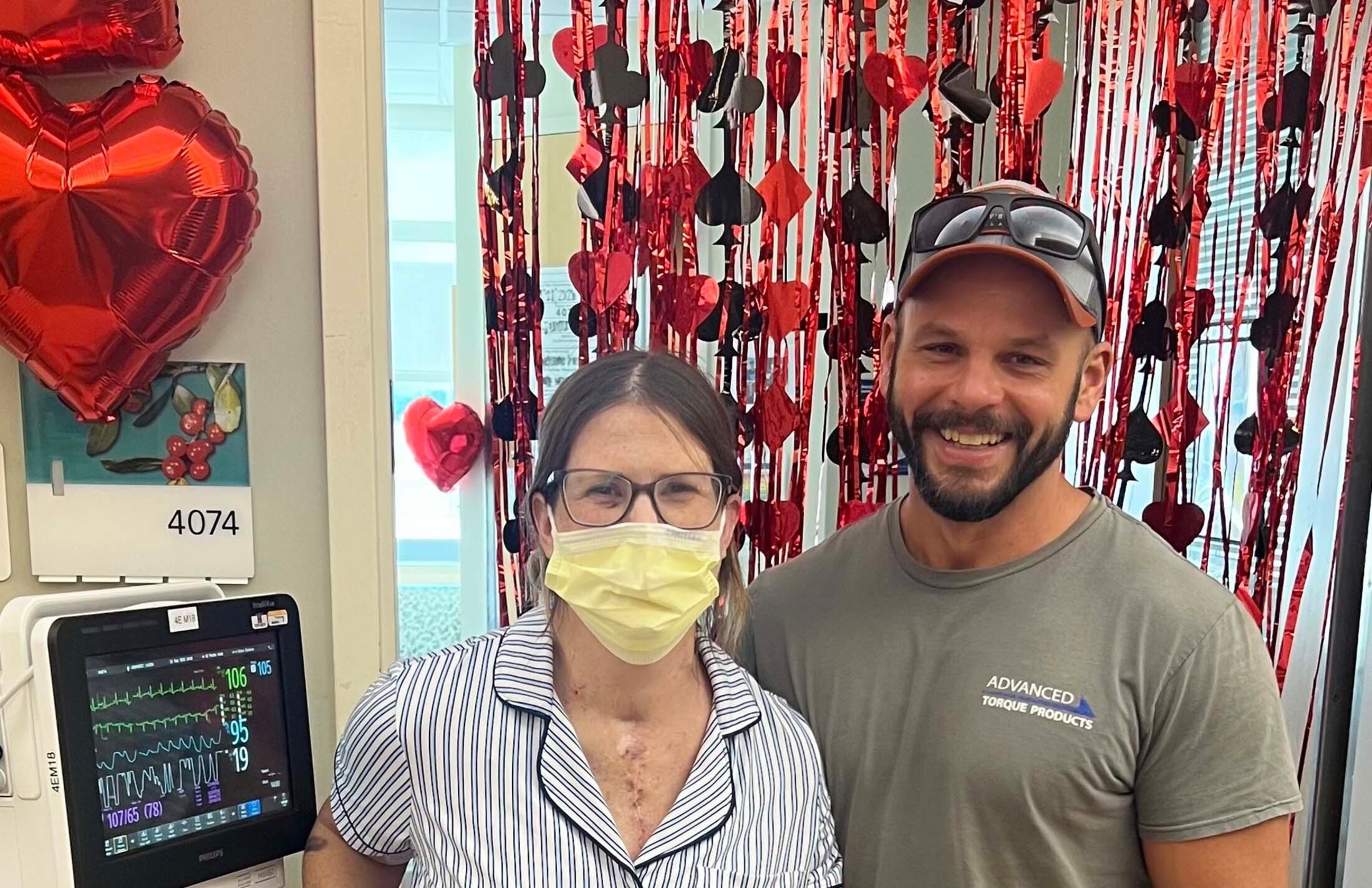 A woman wearing a surgical mask and striped top with a scar on her chest with a smiling man wearing a ball cap stand in front of a curtain of spades, hearts and diamonds. There is a medical device next to the woman.