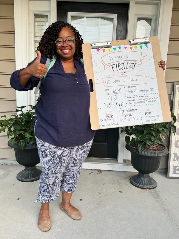 Woman stands in front of her house holding a first day of school sign and giving a thumbs up.